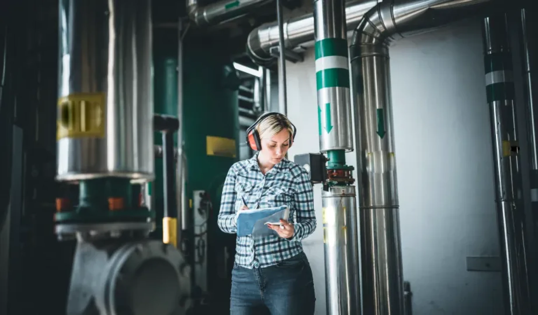 A female engineer wearing ear defenders, looks at a clipboard, in a plant room