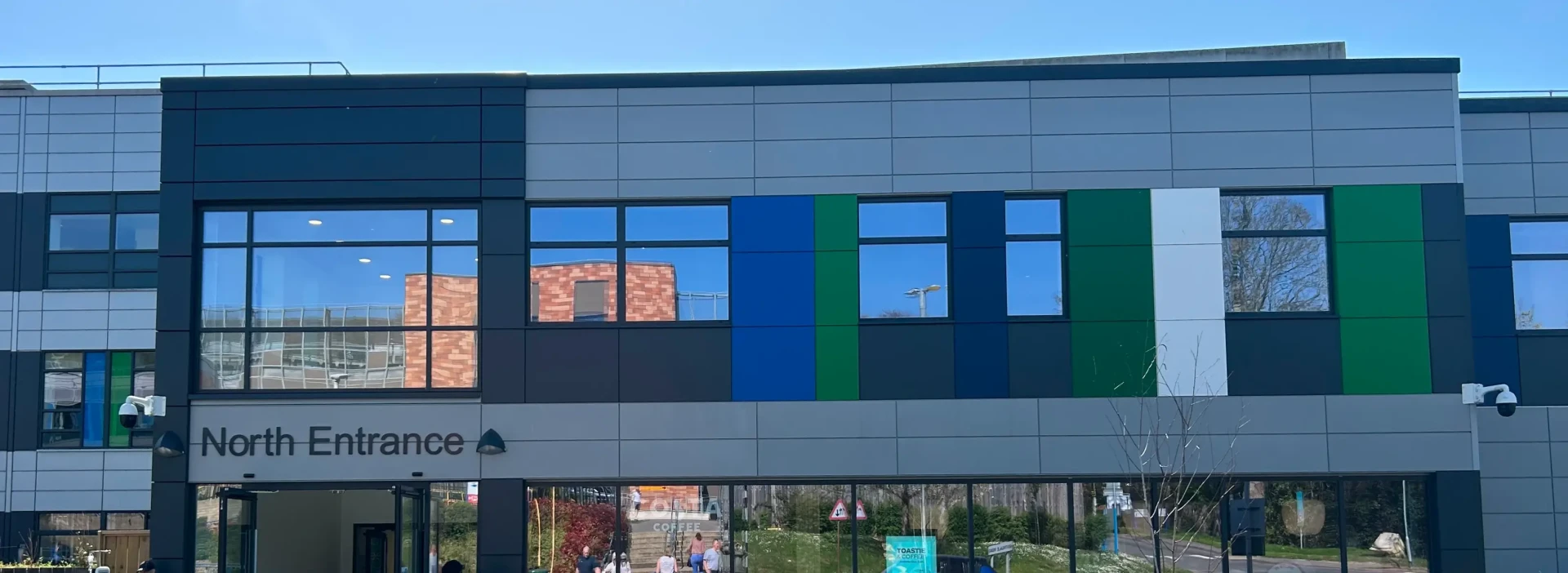 North entrance of a modern hospital building with green and blue panels, and reflective modern windows.