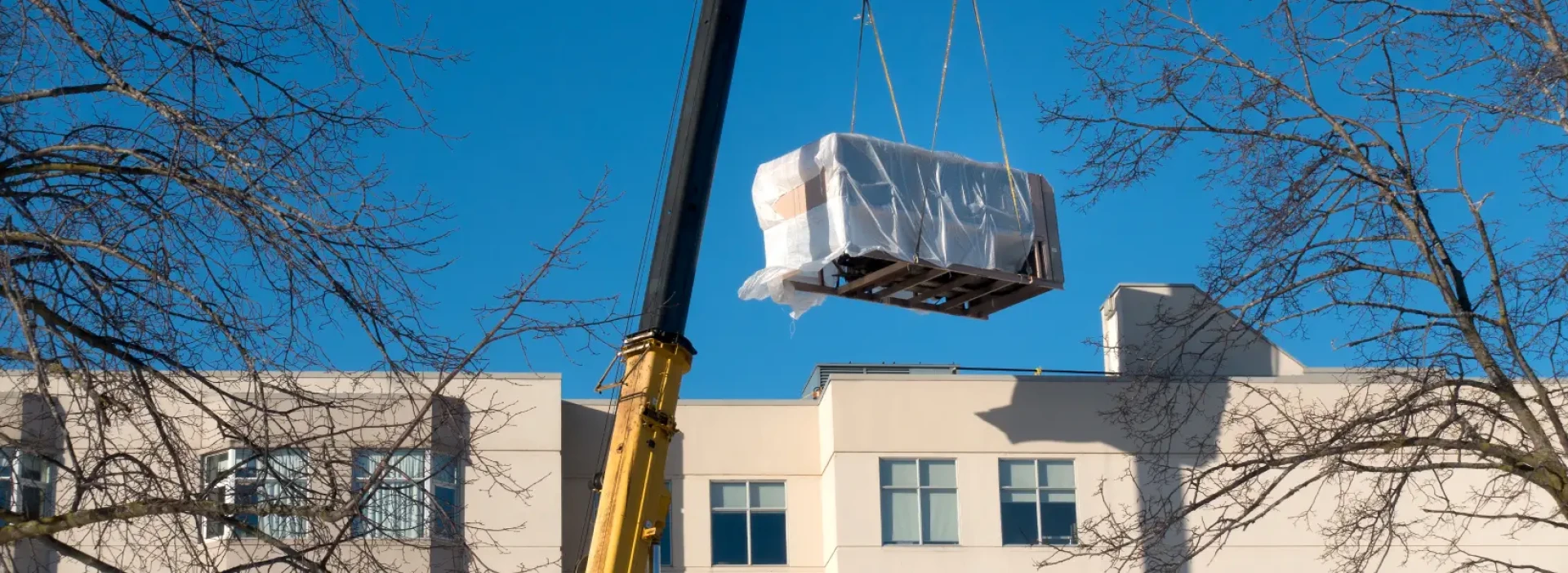 A large industrial crane lifts a heating, ventilation and air conditioning unit onto the roof of an apartment building.