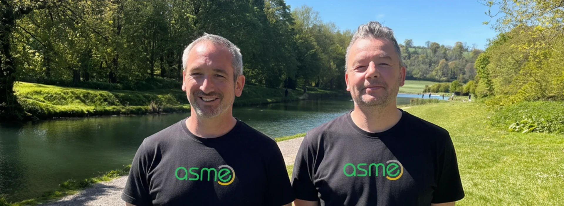 Mark and Andy smiling, wearing asme t shirts, with green trees and a river in the background.