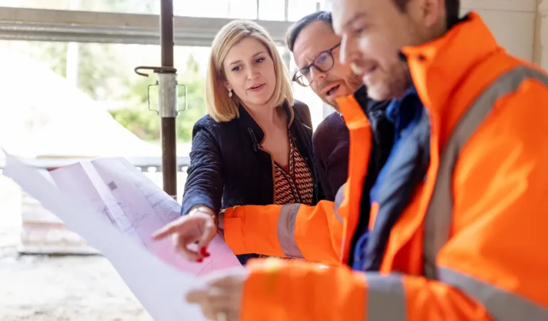 A female in a suit and 2 men in high vis, discuss design plans on a construction site.